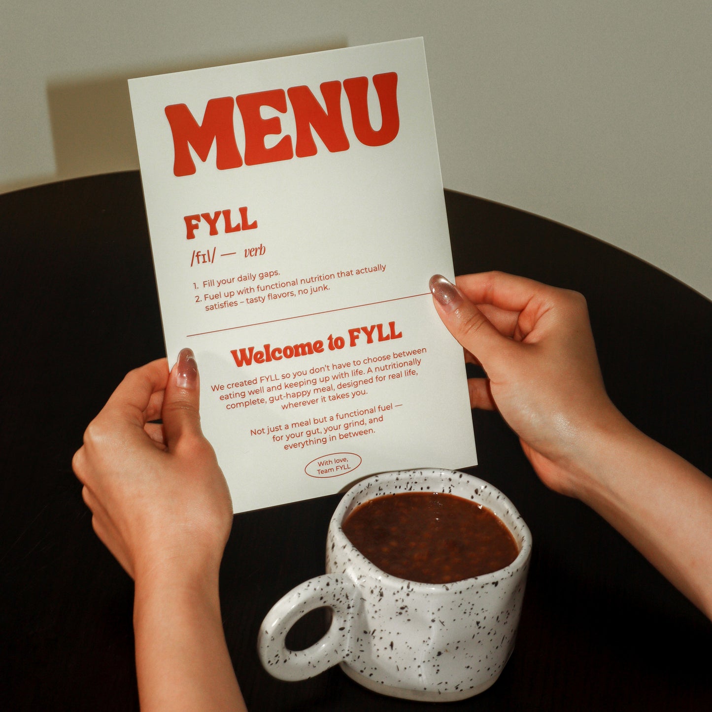 Person holding a 'MENU FYLL' with a cup of coffee on a table